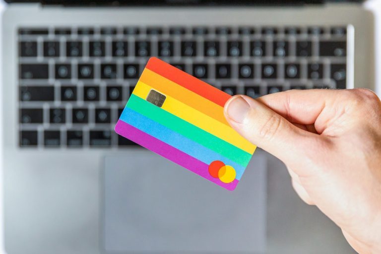 Was kostet das? Eine Hand hält eine Regenbogen-Kreditkarte über einer Laptop-Tastatur.