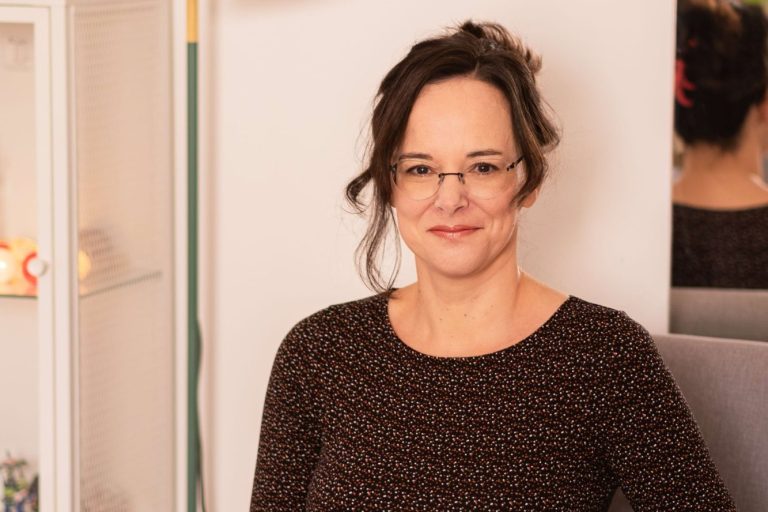 Isabelle Dulleck Frau mit Brille und dunkler Bluse lächelt vor hellen, skandinavischen Möbeln.