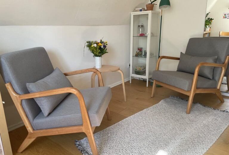 Wo findet die Beratung statt? Zwei moderne Sessel mit Holzgestell, Tisch und Blumen in einem hellen Raum.